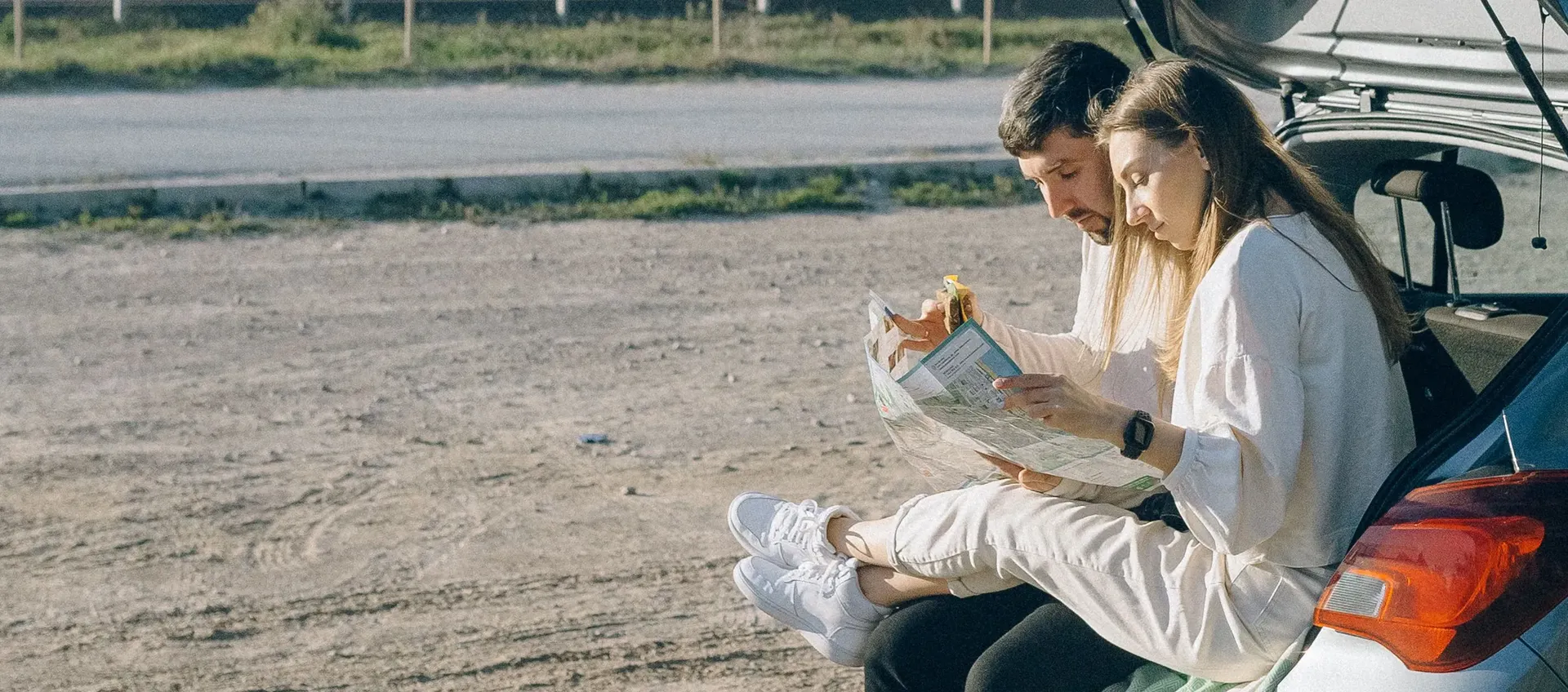 Ein junges Paar sitzt am Rand des offenen Kofferraums eines Kombis, der auf einem Parkplatz neben einer Straße steht, liest Zeitung und isst ein Sandwich.