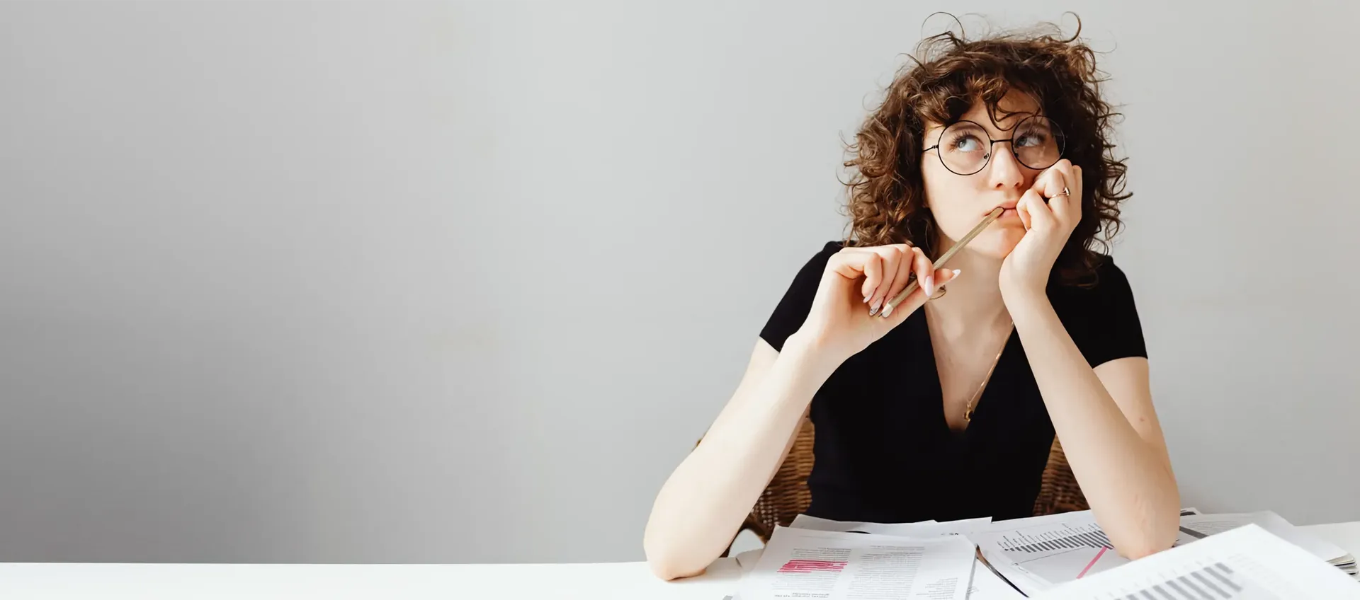 Eine junge Frau sitzt an einem Schreibtisch vor einem Stapel Papierblätter mit Berechnungen, Diagrammen und Texten und denkt nach, während sie in die Ferne blickt.